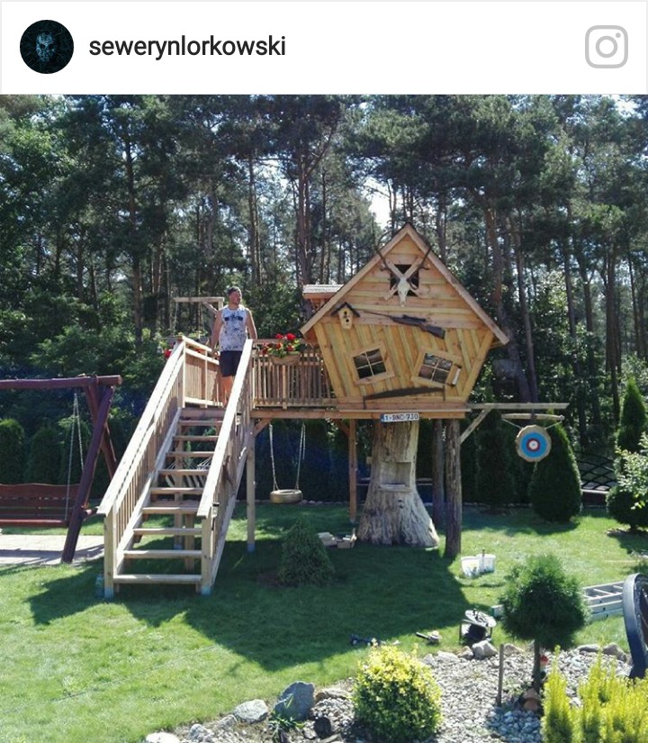 Drewniana konstrukcja domku na drzewie z tarasem, schodami i huśtawką na tle ogrodu z drzewami, ozdobiona czaszką zwierzęcia, imitacją broni i tablicą do strzelania.