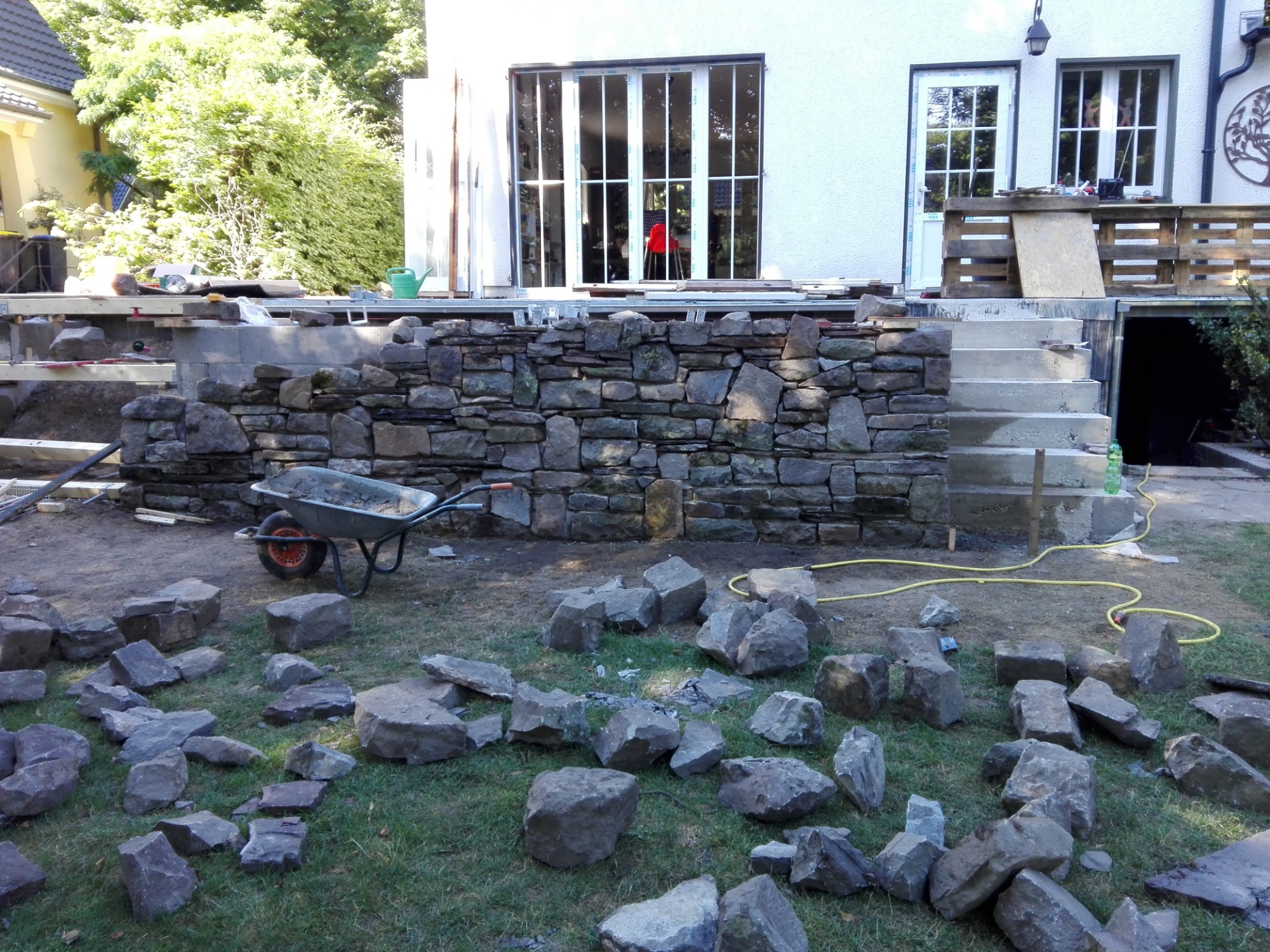 Budowa kamiennej altany: wózek z zaprawą, porozrzucane kamienie na trawie, częściowo ukończony murek z kamienia przed domem z dużymi oknami.