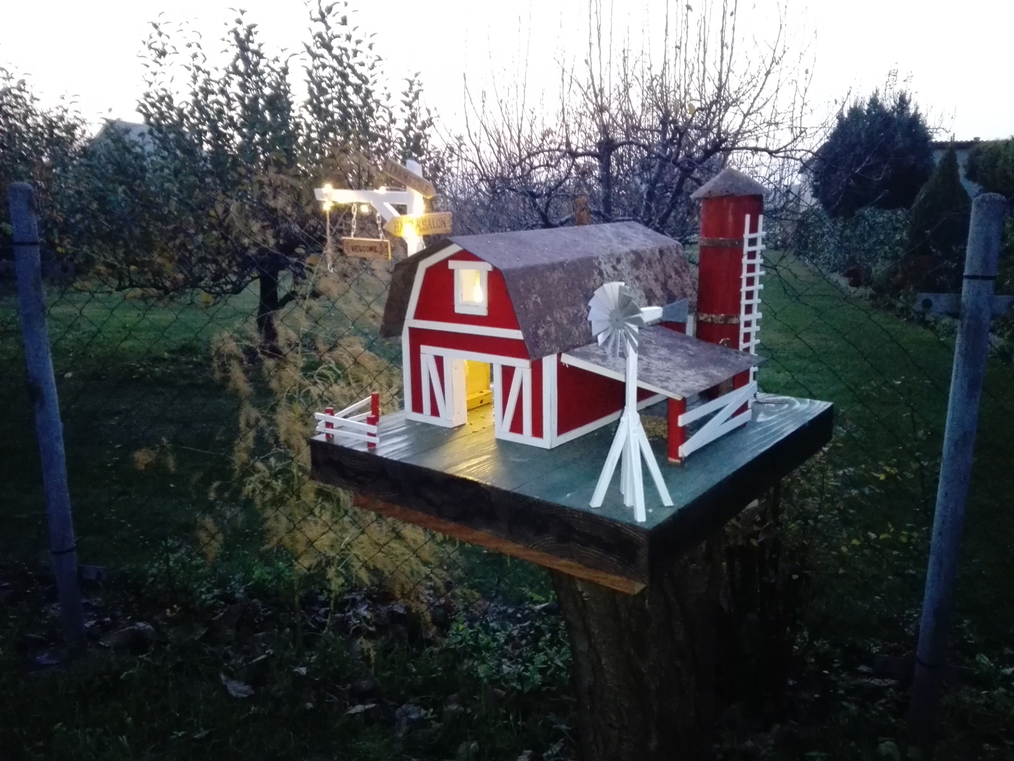 Miniaturowa, podświetlona stodoła z wiatrakiem i wieżą, umieszczona na pniu drzewa w ogrodzie, za siatką.