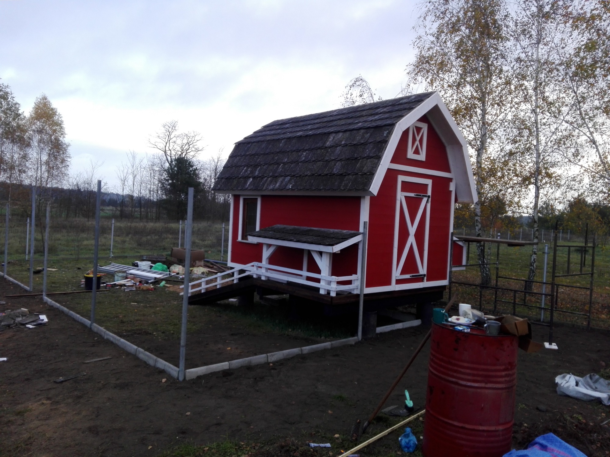 Czerwona, drewniana altana w stylu stodoły z białymi detalami, częściowo otoczona metalowym ogrodzeniem, na tle jesiennego krajobrazu z drzewami.