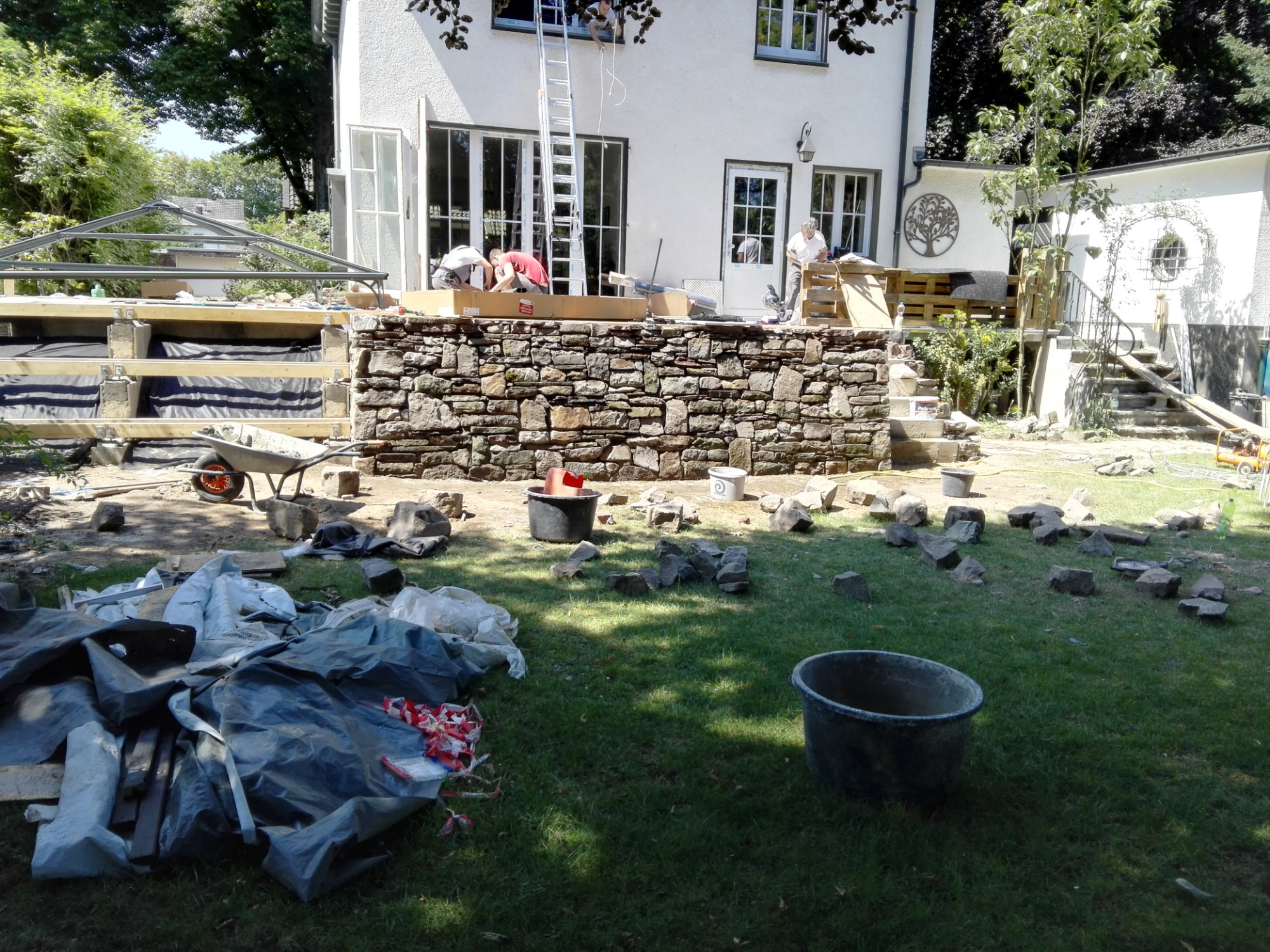 Budowa altany: murowana podstawa z kamienia, widoczna konstrukcja dachu altany, pracownicy na drabinie przy budynku, przygotowane materiały budowlane na trawniku.