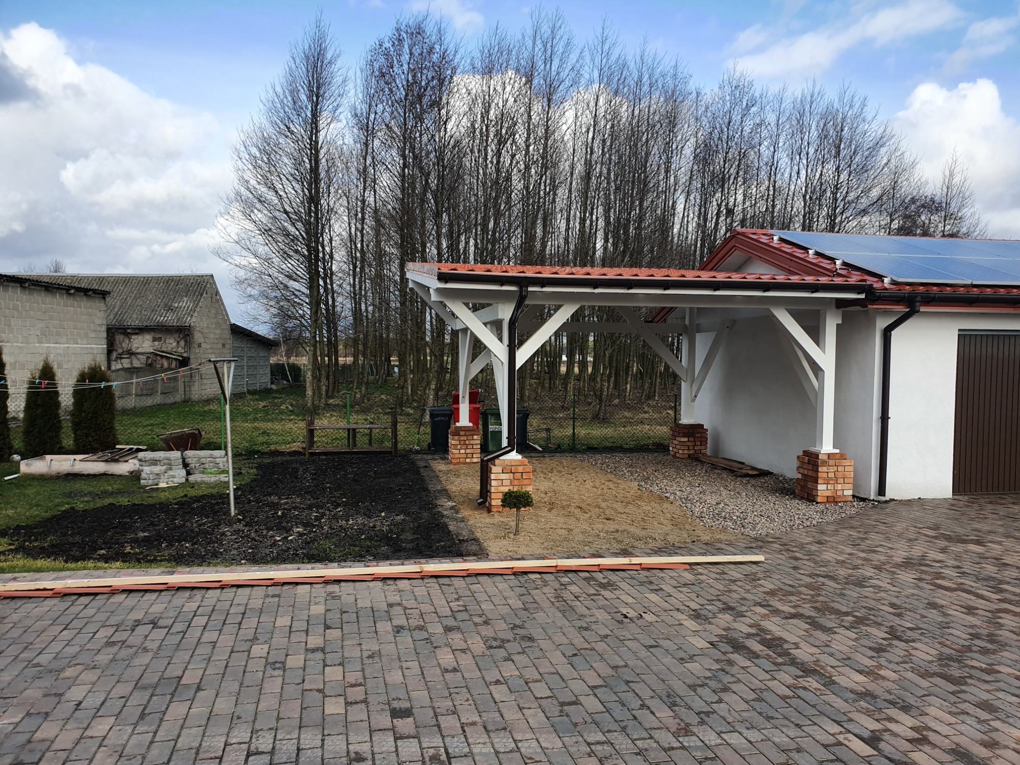 Biała konstrukcja z czerwonym dachem, wsparta na ceglanych filarach, przylegająca do białego budynku z panelami słonecznymi, widoczna z brukowanego podjazdu, z przygotowanym terenem pod nasadzenia...