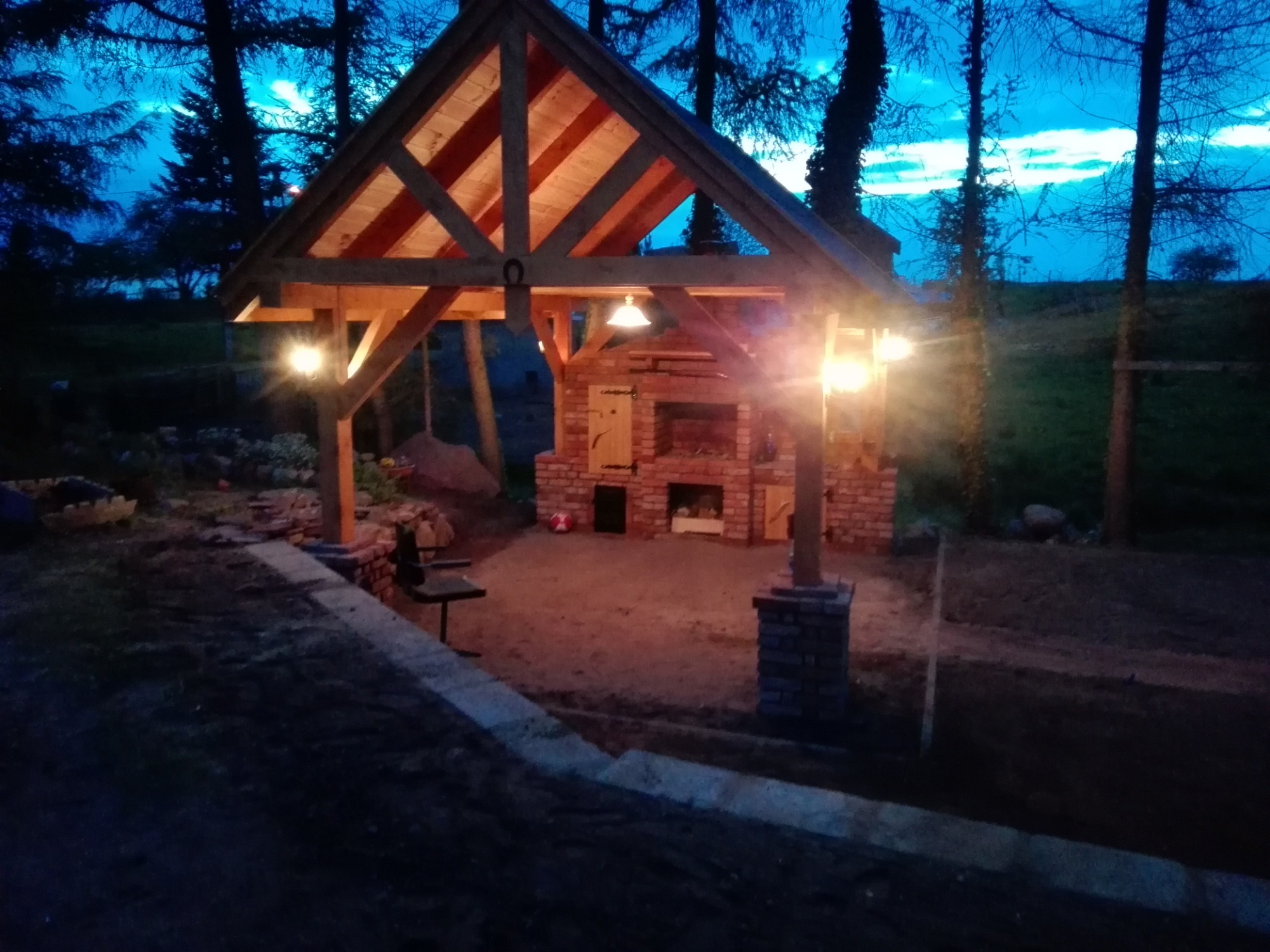 Drewniana konstrukcja z ceglanym piecem i oświetleniem w ogrodzie o zmierzchu, widoczny podkowy na konstrukcji.