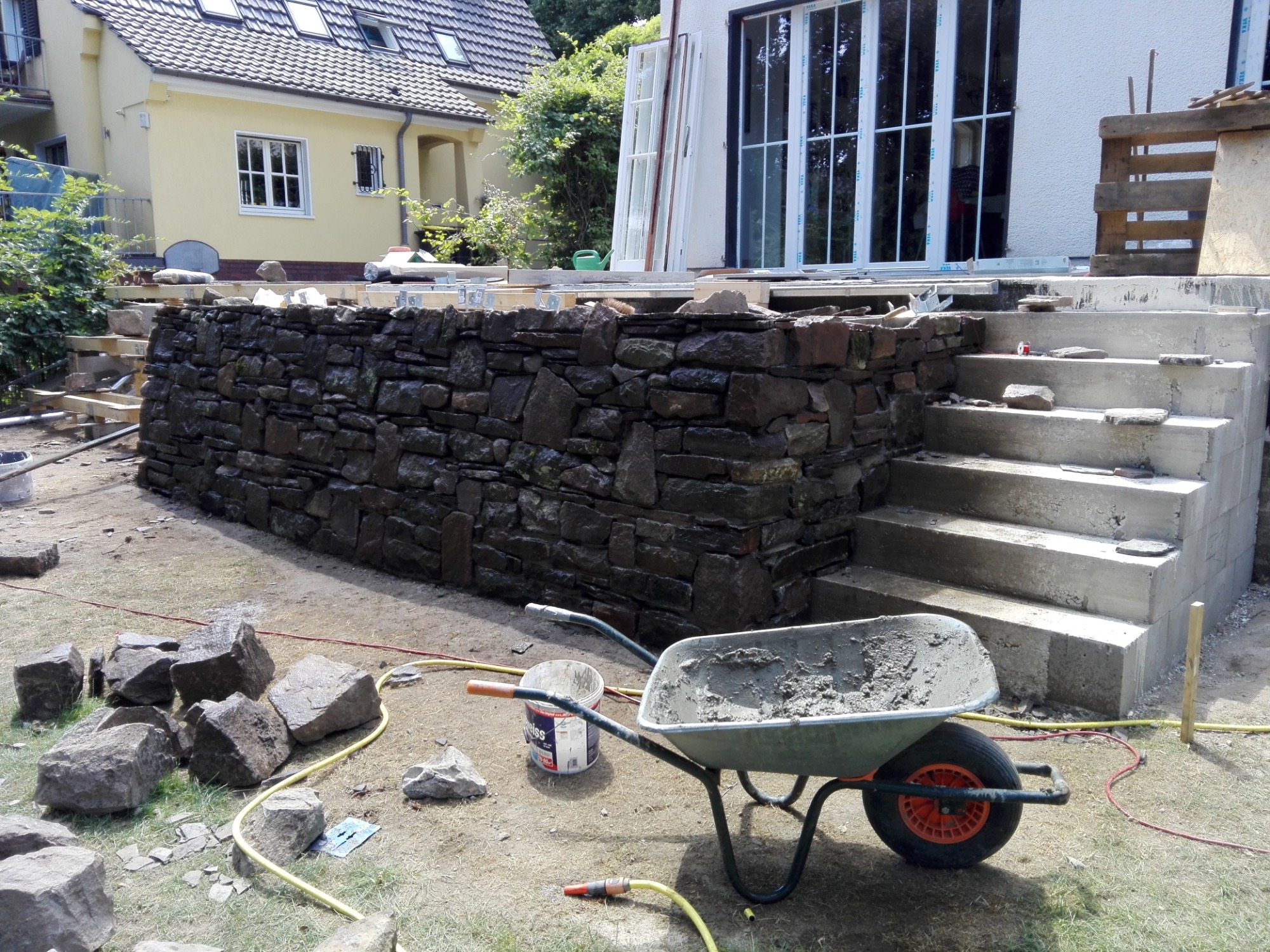 Budowa kamiennej ścianki oporowej przed domem, widoczna taczka z zaprawą, ułożone kamienie, schody wejściowe do budynku w trakcie budowy.