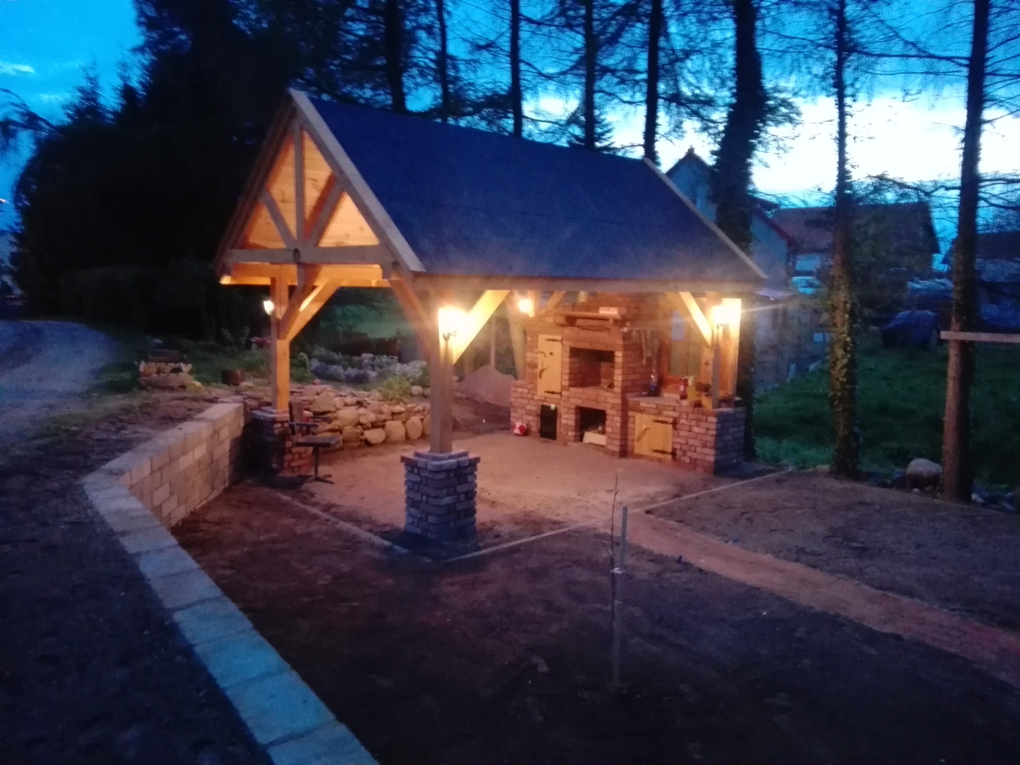 Zadaszona konstrukcja z drewna i cegły z oświetleniem, z piecem/grillem murowanym wewnątrz, otoczona ziemią i drzewami w wieczornym oświetleniu.