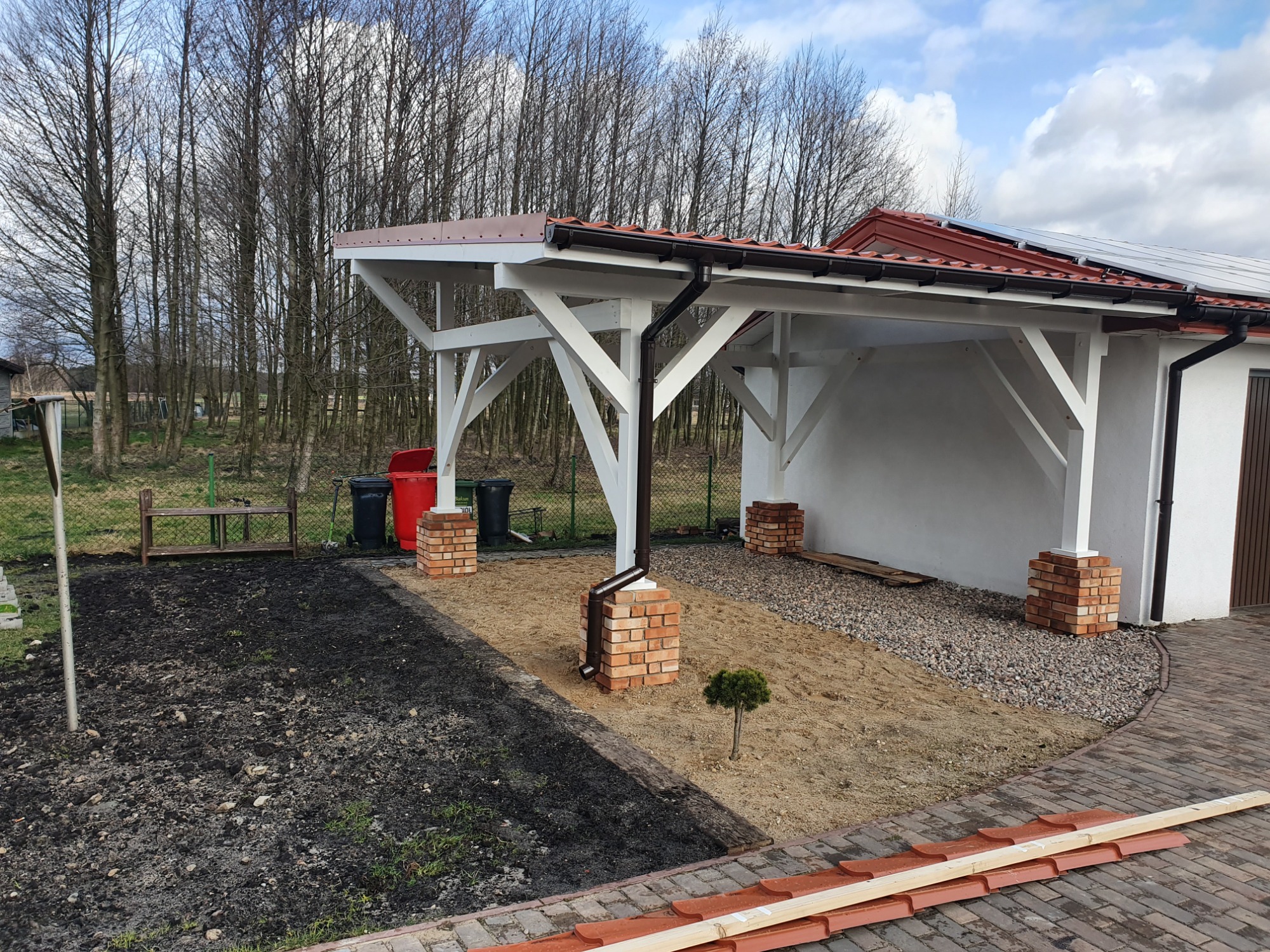 Biała konstrukcja z brązowym dachem, wsparta na ceglanych filarach, z widocznymi panelami słonecznymi na dachu budynku w tle, teren przygotowany pod nasadzenia.