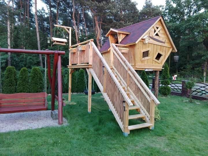 Drewniany domek na drzewie z ukośnymi oknami i schodami, obok huśtawka na tle zielonego ogrodu i drzew.