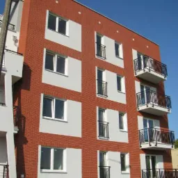 Nowoczesny, czteropiętrowy budynek mieszkalny z czerwonej cegły z białymi elementami elewacji i czarnymi balkonami, widziany z dołu pod kątem.