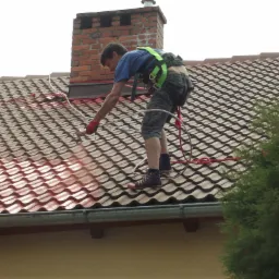 Pracownik w uprzęży, malujący czerwoną farbą dachówkę na dachu domu za pomocą pistoletu natryskowego, widoczny komin z cegły i fragment iglaka.