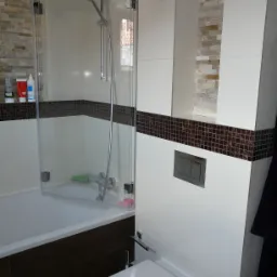 Nowoczesna łazienka z białymi płytkami, mozaikowym pasem, kabiną nawannową z przezroczystego szkła i toaletą typu Geberit.