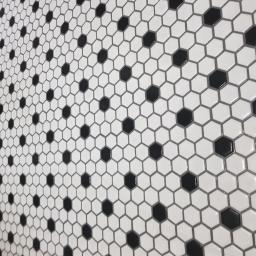 W.B-wykonczenia - Białe, heksagonalne płytki z czarnymi akcentami ułożone na ścianie, widoczne pod kątem, z delikatnym połyskiem.