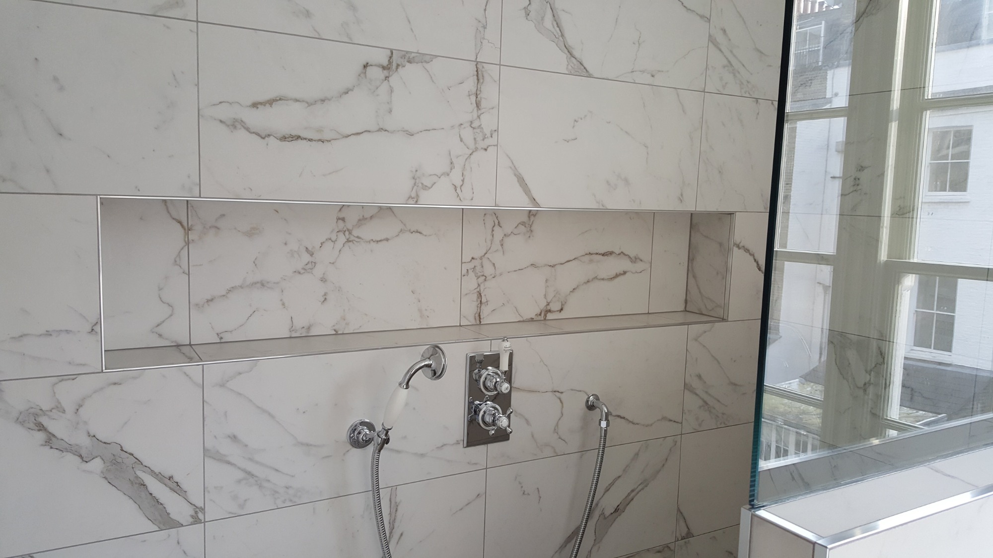 Wykończona kabina prysznicowa z białymi płytkami imitującymi marmur, wnęka w ścianie wykończona listwami, chromowana bateria z dwoma kurkami oraz słuchawką prysznicową na wężu, widok przez szybę...