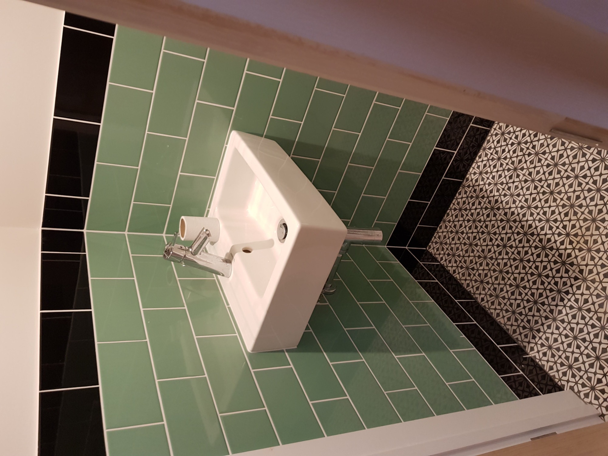 Nowoczesna łazienka z zielonymi, podłużnymi płytkami na ścianie, białą umywalką i czarno-białą mozaiką na podłodze, widziana z góry pod kątem.