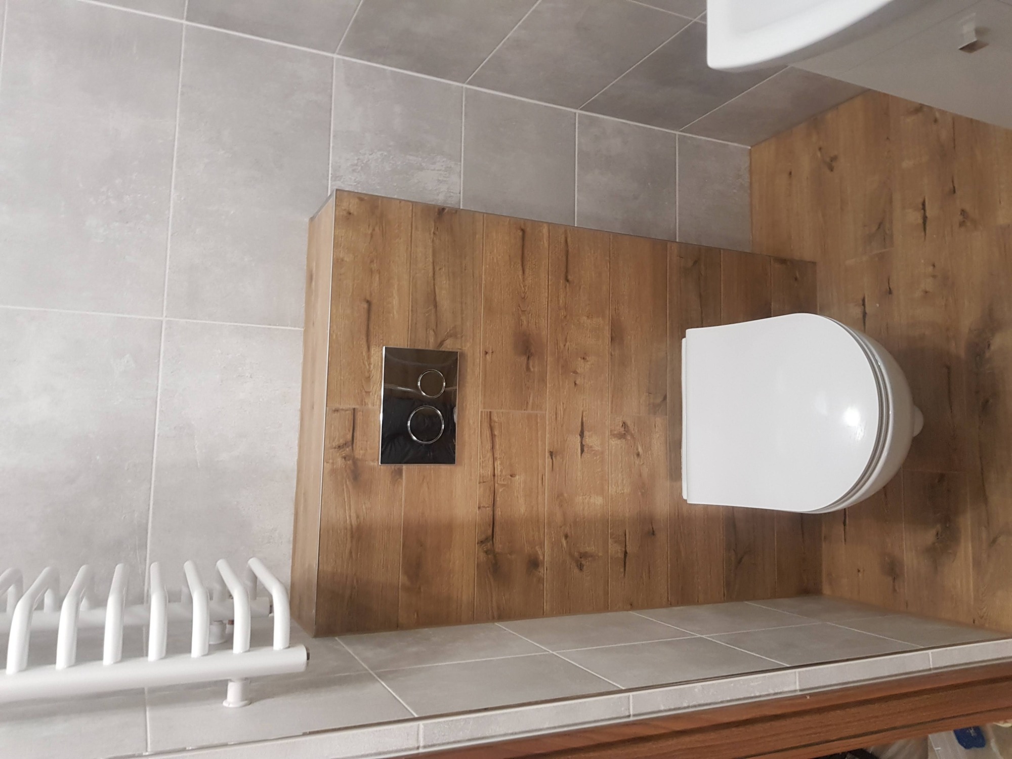 Wykończona łazienka z szarymi płytkami na ścianach i podłodze, drewnopodobną zabudową ściany za toaletą, chromowanym przyciskiem spłuczki i białym grzejnikiem drabinkowym.