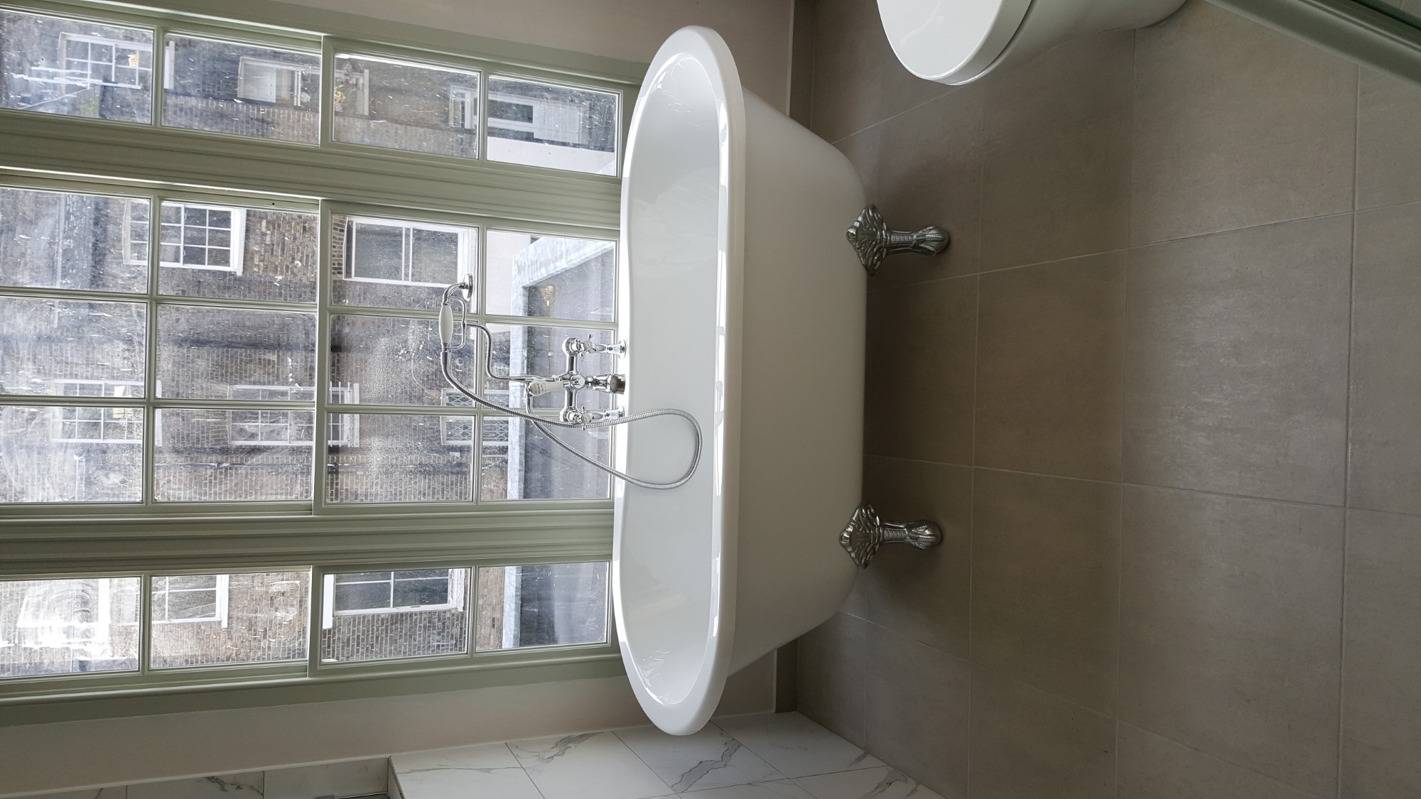 Wolnostojąca, biała wanna na ozdobnych nóżkach w łazience z dużym oknem z widokiem na budynek z cegły.