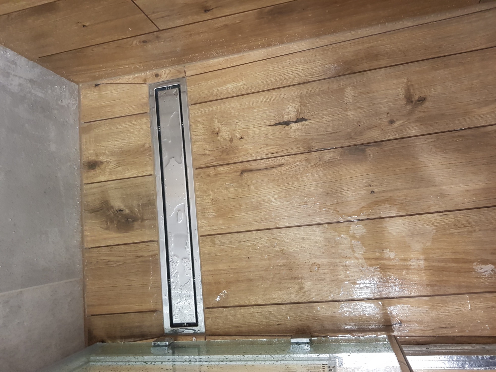 Ukończona łazienka z odpływem liniowym w podłodze wyłożonej panelami imitującymi drewno i szarą płytką na ścianie, widoczne krople wody.