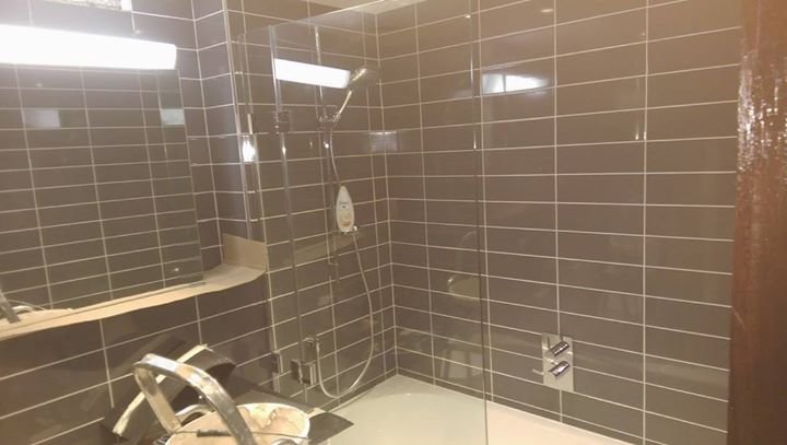 Nowoczesna kabina prysznicowa z szarymi, podłużnymi płytkami, chromowaną armaturą i szklaną ścianą, widok z perspektywy umywalki z metalowym koszem.