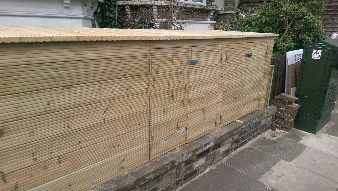 Drewniana zabudowa z ryflowanych desek, prawdopodobnie na śmietniki, z trzema parami drzwiczek, zabezpieczonych metalowymi skobelkami, zamontowana na murku z cegieł