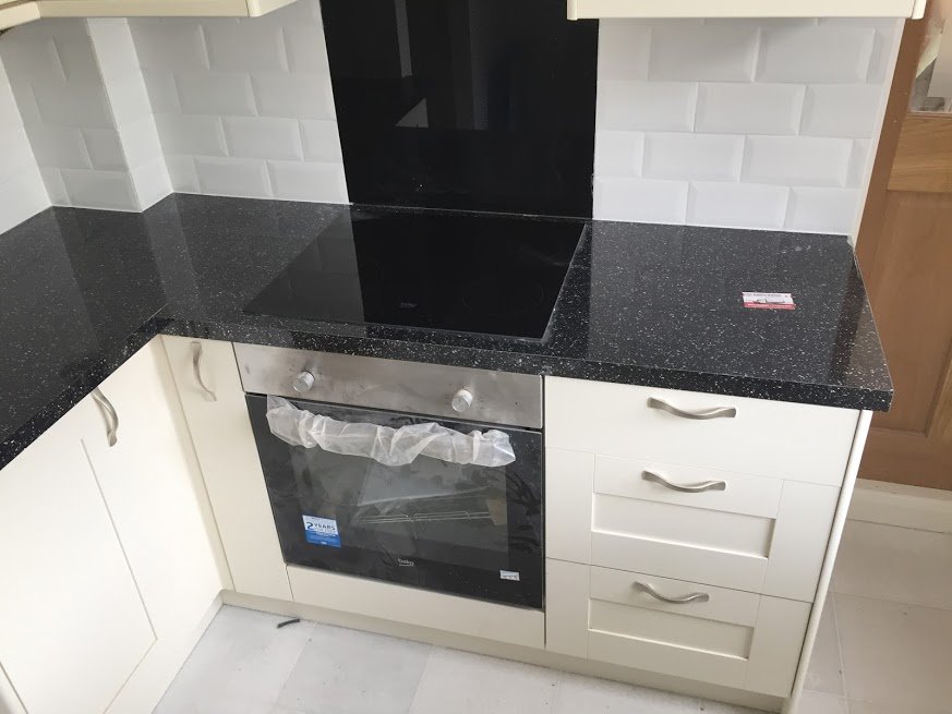 Kremowa zabudowa kuchenna z czarnym, błyszczącym blatem, płytą indukcyjną i piekarnikiem z folią ochronną na drzwiczkach, białe płytki na ścianie.