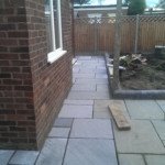 Ułożony chodnik z płyt kamiennych wzdłuż ściany budynku z cegły klinkierowej, widoczne ogrodzenie drewniane i fragment ogrodu.