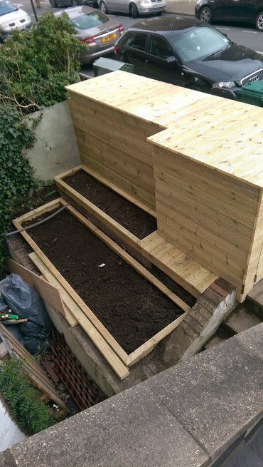 Dwa drewniane podwyższone kwietniki wypełnione ziemią, zabudowane drewnianą ścianą, widok z góry na tle ulicy z samochodami.