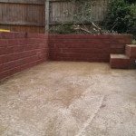 Ceglana ścianka oporowa z dwoma stopniami na betonowym podłożu, otoczona drewnianym płotem.