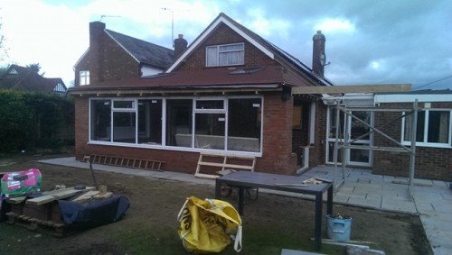 Rozbudowa domu z cegły z dużymi oknami i przygotowanym zadaszeniem tarasu, widoczne materiały budowlane i rusztowania.