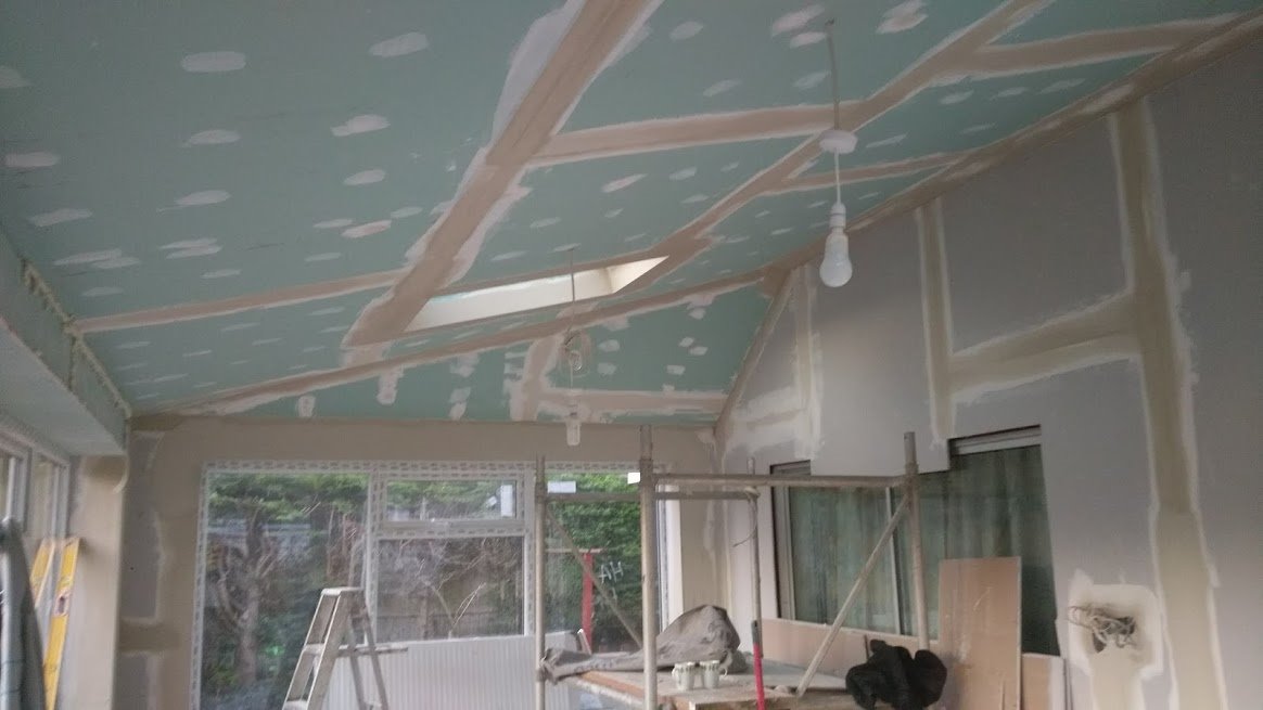 Wnętrze w trakcie prac wykończeniowych: zamontowane płyty gipsowo-kartonowe na suficie ze szpachlowanymi łączeniami, widoczne okna i drabina w pomieszczeniu.