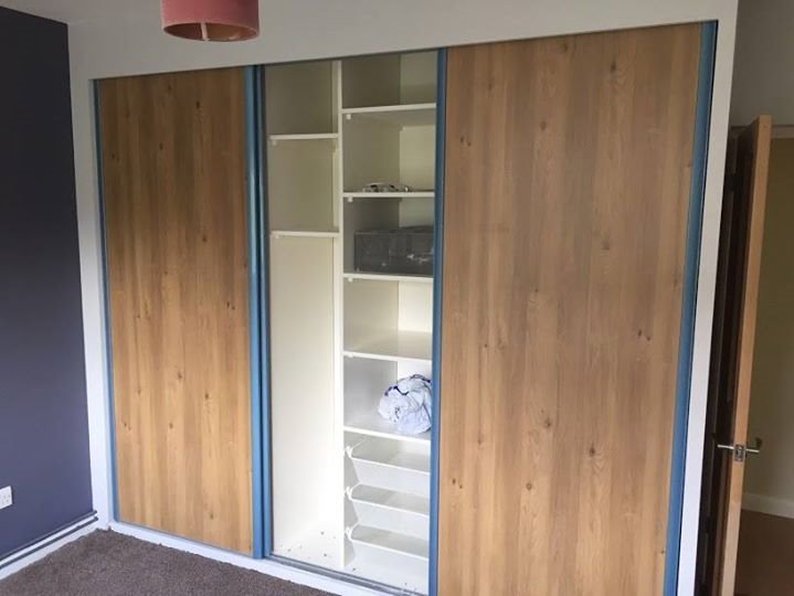 Wbudowana szafa z przesuwnymi drzwiami o fakturze jasnego drewna, z niebieskimi obramowaniami, częściowo odsłaniająca białe półki z zawartością w pokoju z szarą ścianą i brązową wykładziną.