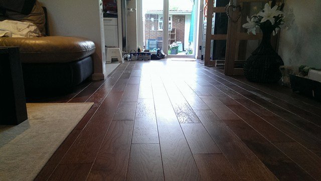 Błyszcząca, świeżo położona podłoga z paneli drewnianych w salonie, z widocznym fragmentem sofy i wyjściem na ogród w tle.