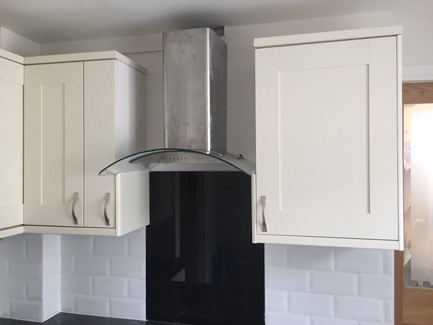 Białe szafki kuchenne z frezowanymi frontami, srebrny okap z czarnym panelem szklanym, białe kafelki typu metro i czarny blat.