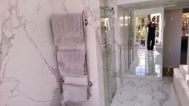 Nowoczesna łazienka z marmurowymi ścianami i podłogą, szklana kabina prysznicowa, grzejnik drabinkowy na ręczniki z trzema złożonymi ręcznikami, osoba robiąca zdjęcie w tle.