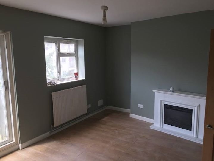 Pomalowane na szaro wnętrze pokoju z parkietem, białym kominkiem elektrycznym i grzejnikiem pod oknem. Widoczne gniazdka elektryczne na ścianie.