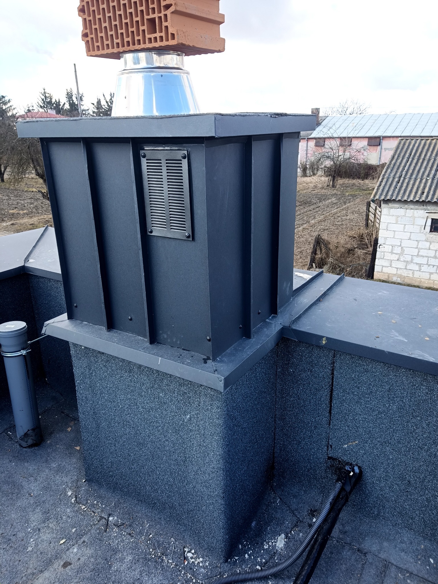 Nowoczesny komin wentylacyjny z czarną obudową blacharską, zakończony ceglanym elementem, widoczny na tle dachu pokrytego papą.