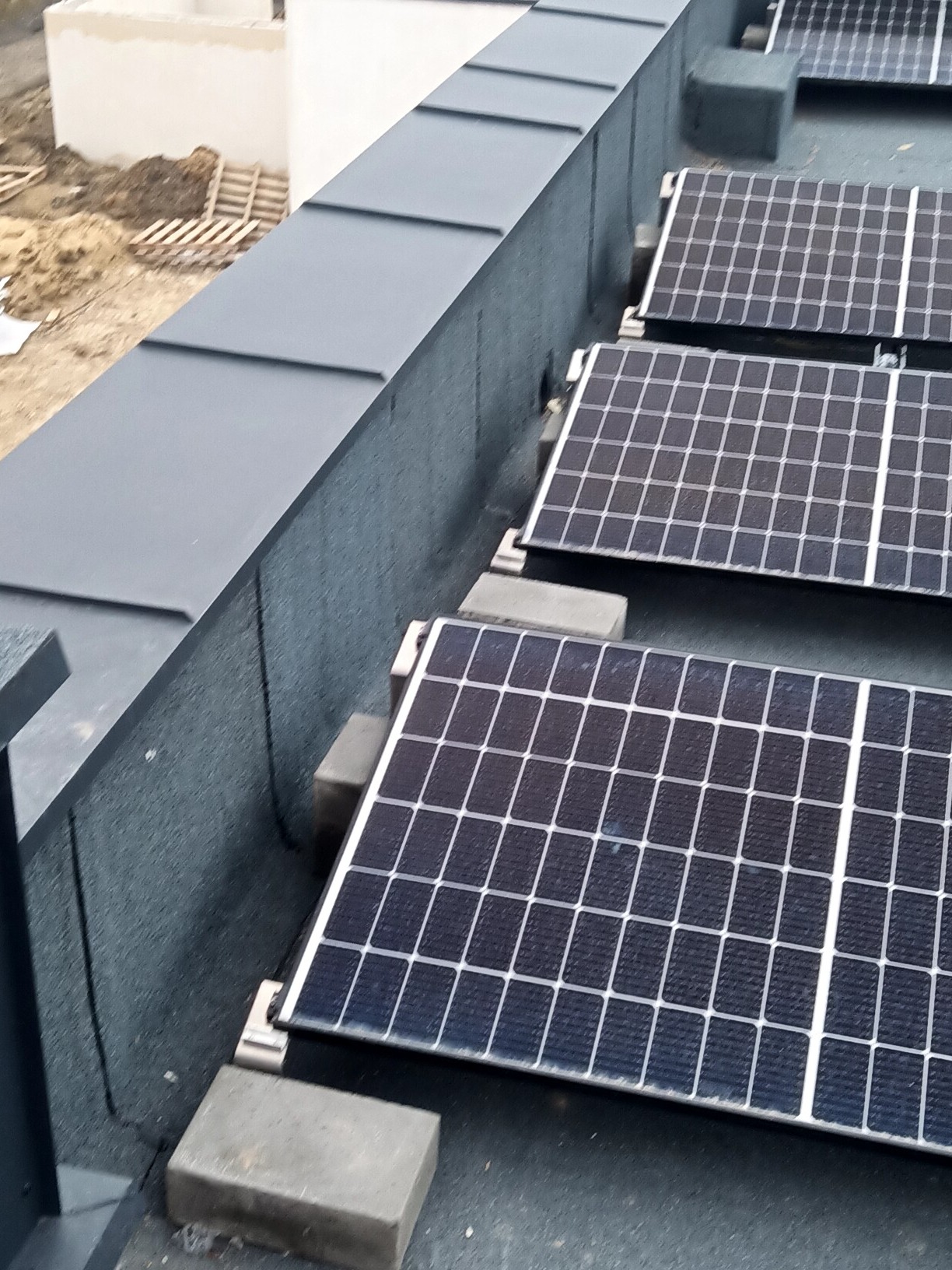 Panele słoneczne zamontowane na płaskim dachu z obróbką blacharską, widoczne betonowe podstawy pod panele.