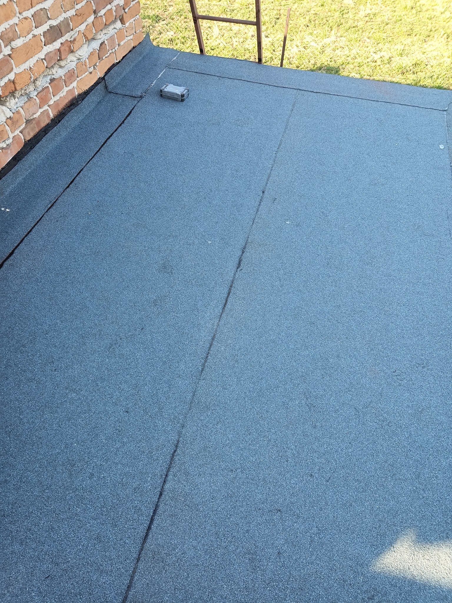 Płaski dach pokryty szarą papą termozgrzewalną z widocznym łączeniem, fragment ściany z czerwonej cegły oraz drabina w tle.