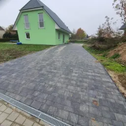 Ułożona szara kostka brukowa prowadząca do zielonego domu z szarym dachem, widoczny drenaż liniowy na pierwszym planie oraz częściowo przygotowany teren po prawej stronie.