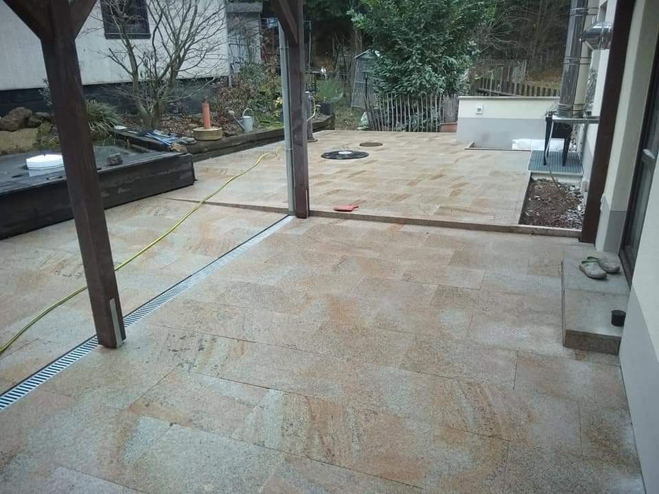 Wykończony taras z beżowo-szarych płyt kamiennych, z odpływem liniowym, drewnianą pergolą i widocznymi butami przy wejściu do budynku.