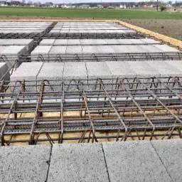 Zbrojenie fundamentów z bloczków betonowych ułożone na szalunku, widok z góry na tle zielonego pola i zabudowań.