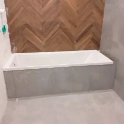 Wanna obudowana szarą płytką, z drewnopodobną okładziną ściany ułożoną w jodełkę w tle, widoczne białe płytki oraz instalacja sanitarna na ścianie po lewej.