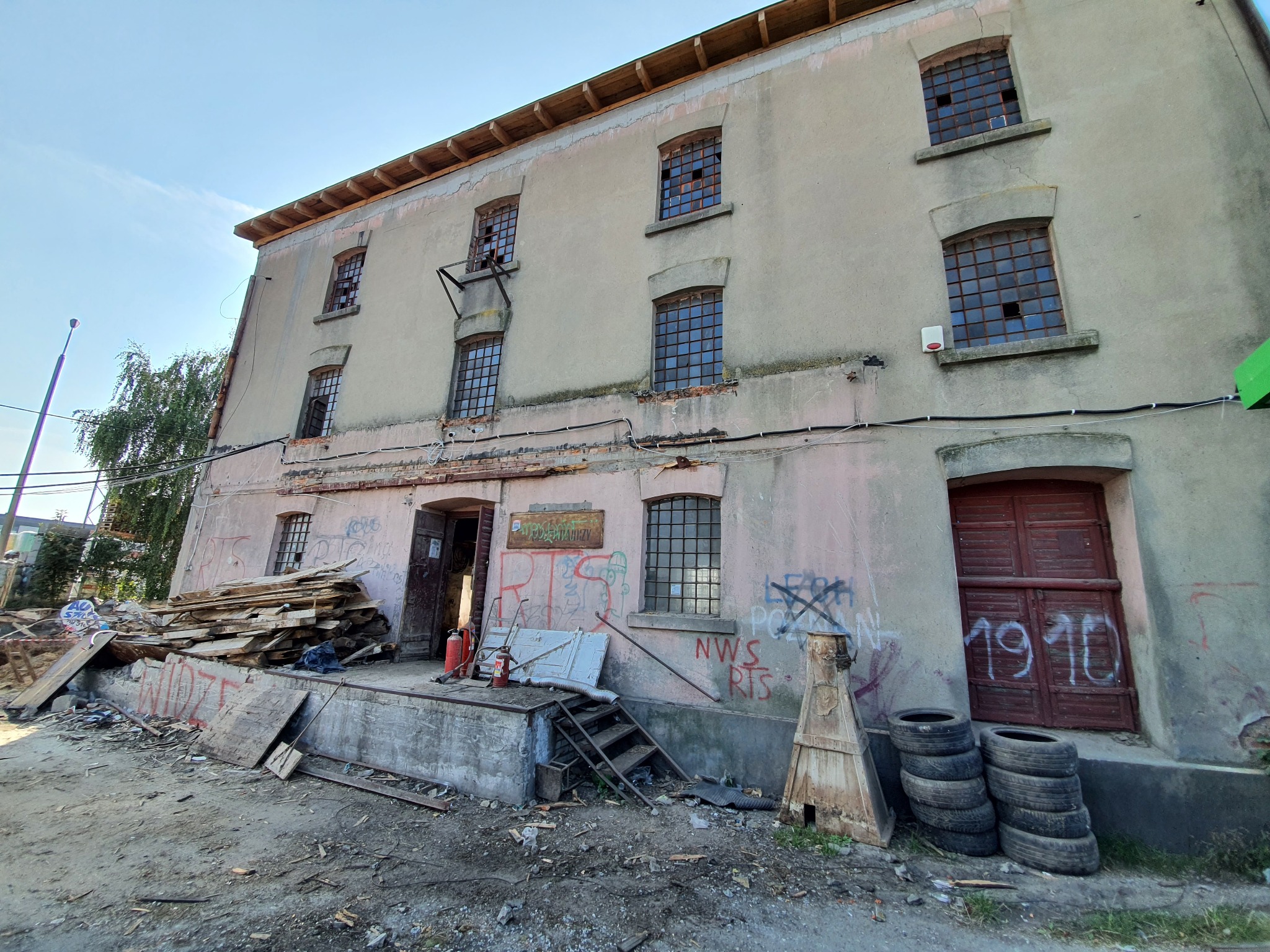 Stary, zaniedbany budynek z wybitymi oknami, stertą desek i graffiti na ścianach, prawdopodobnie przygotowywany do rozbudowy lub remontu.