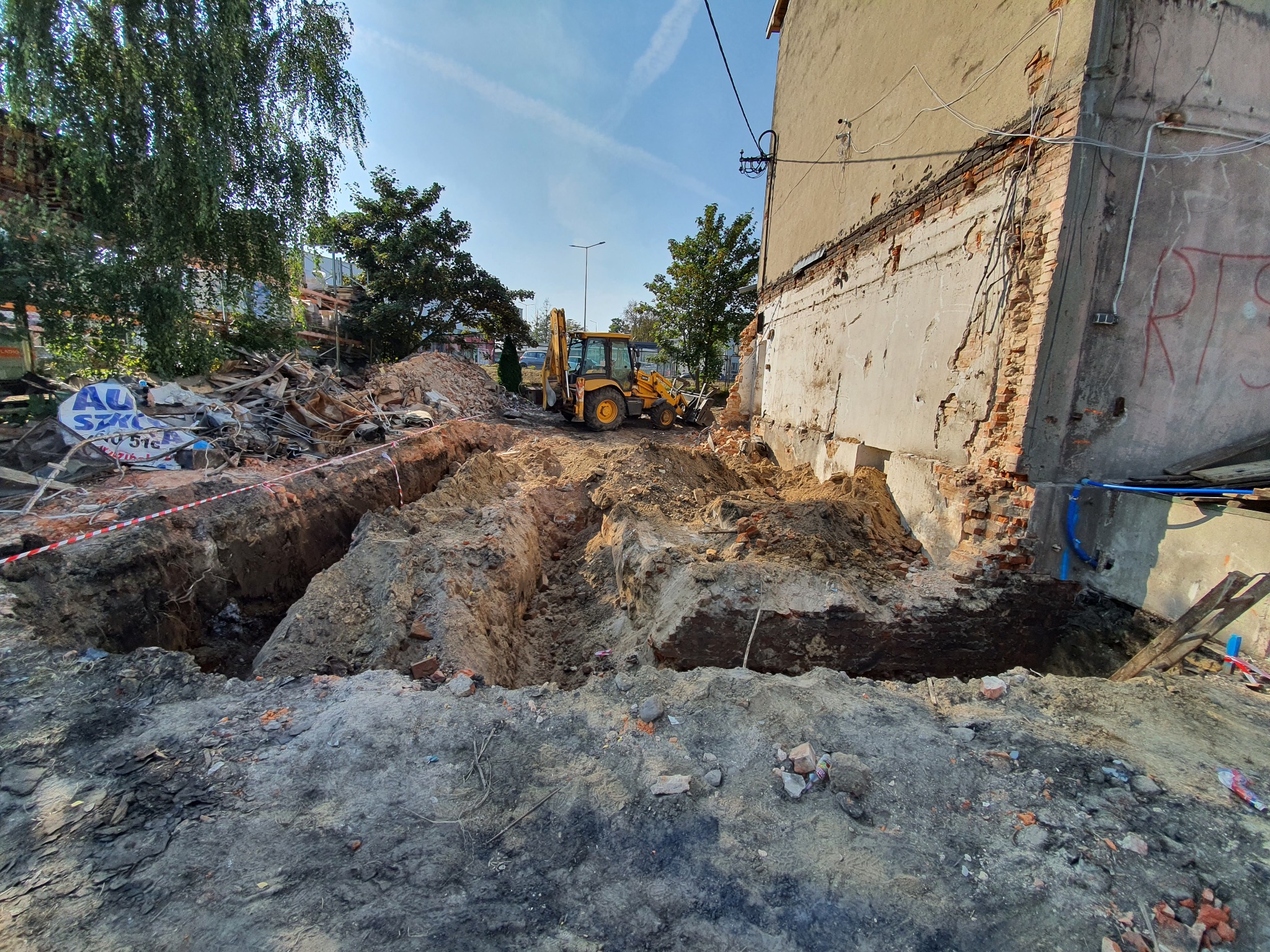 Wykop fundamentowy przy budynku z odsłoniętą cegłą, z widoczną koparką w tle i gruzem po rozbiórce.