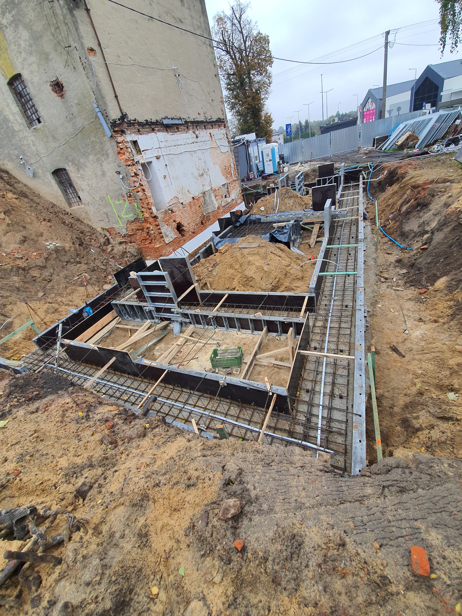 Przygotowanie fundamentów: szalunki i zbrojenie na placu budowy, widok z góry, w tle budynek z cegły.