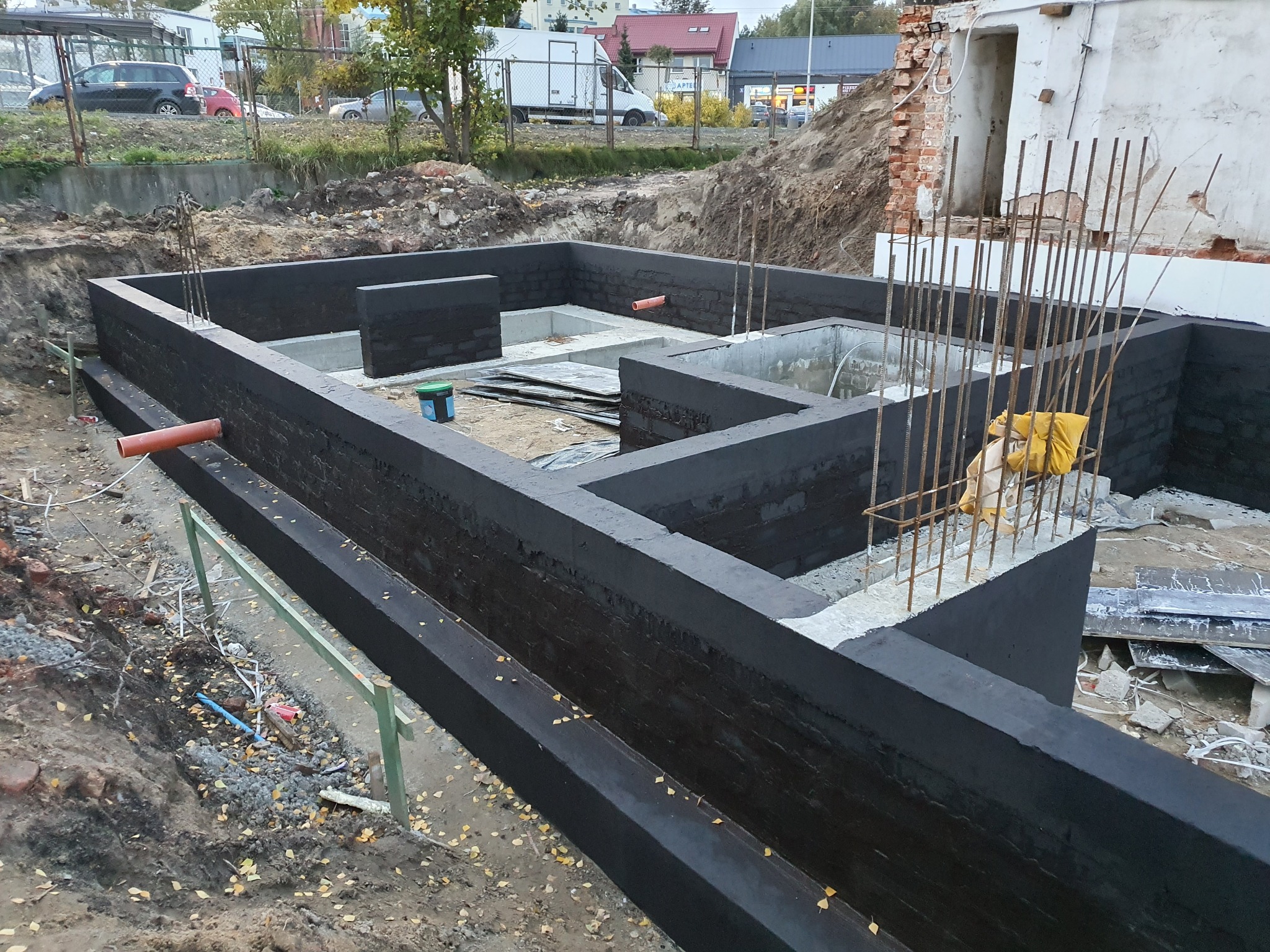 Wykopane fundamenty domu z hydroizolacją, z wystającymi prętami zbrojeniowymi, widoczna puszka farby i pozostałości starego budynku w tle.
