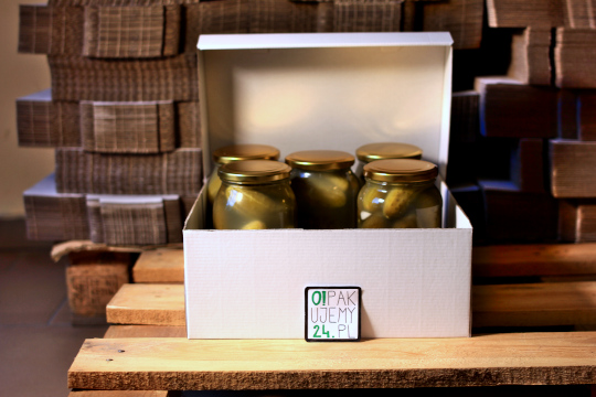 Białe pudełko z pięcioma słoikami ogórków konserwowych na drewnianej palecie, z logo firmy na pudełku i stosem kartonów w tle.