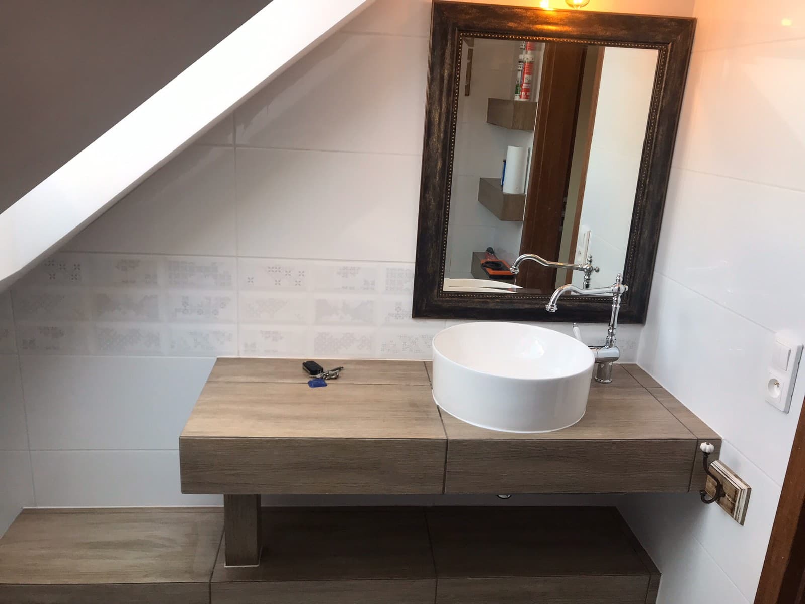 Nowoczesna łazienka na poddaszu: biała umywalka nablatowa, bateria w stylu retro, lustro w ciemnej ramie z oświetleniem, płytki ścienne z delikatnym wzorem, klucze leżące na blacie.