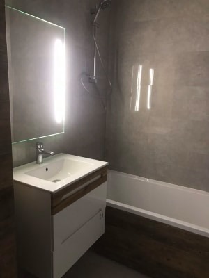 Nowoczesna łazienka z szarymi płytkami imitującymi beton, podświetlanym lustrem nad białą szafką z umywalką i wanną z zabudową z paneli drewnopodobnych.