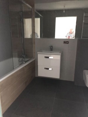 Wykończona łazienka z ciemną podłogą, jasnymi płytkami imitującymi drewno na obudowie wanny, białą szafką pod umywalką z dwoma szufladami i lustrem, w którym odbija się osoba.