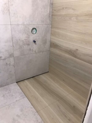 Ułożone płytki imitujące drewno na podłodze i ścianie, z szarymi płytkami na ścianie w tle, widoczny otwór na instalację wodną