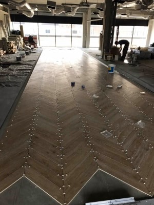 Układanie parkietu w jodełkę z użyciem krzyżyków dystansowych w przestronnym wnętrzu, widoczne puszki z klejem i pracownik w tle.